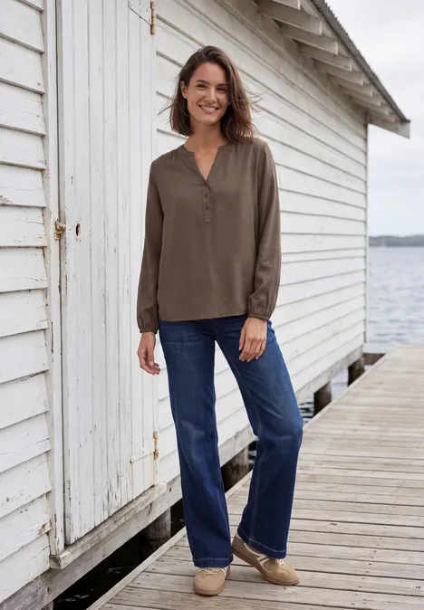 Eine lächelnde Frau in olivgrüner Bluse und Jeans steht auf einem Holzsteg neben einem weißen Bootshaus.