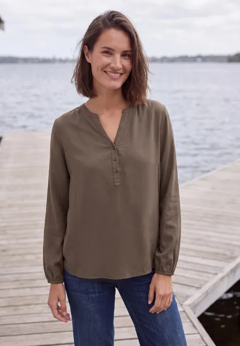 Eine lächelnde Frau steht auf einem Steg am Wasser, trägt eine olivgrüne Bluse und blaue Jeans.