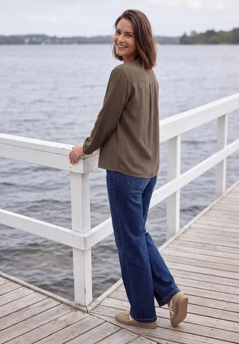 Frau in grüner Jacke und blauer Jeans steht auf einem Steg am Wasser und blickt lächelnd über die Schulter.