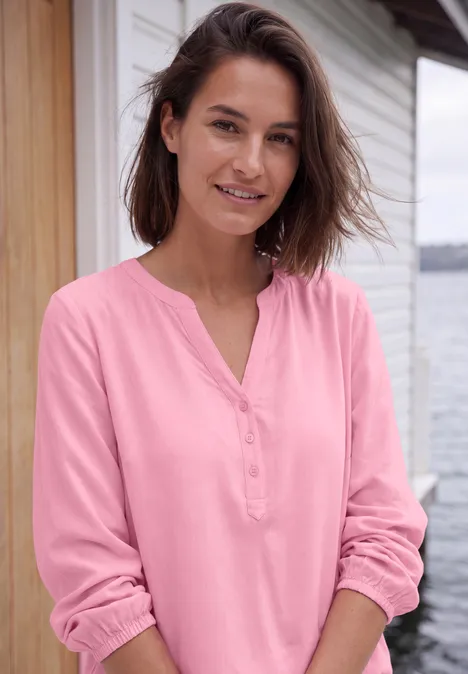 Eine lächelnde Frau in rosa Bluse steht vor einem Haus und blickt entspannt in die Kamera.