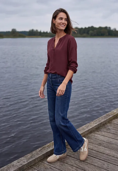 Eine Frau steht entspannt auf einem Steg am See, trägt eine bordeauxrote Bluse, Jeans und helle Schuhe.