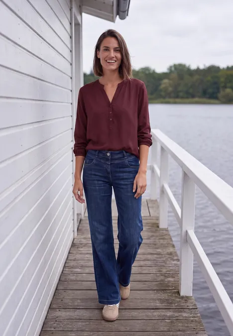 Frau in weinroter Bluse und Jeans steht auf Steg neben weißem Haus am See und lächelt in die Kamera.