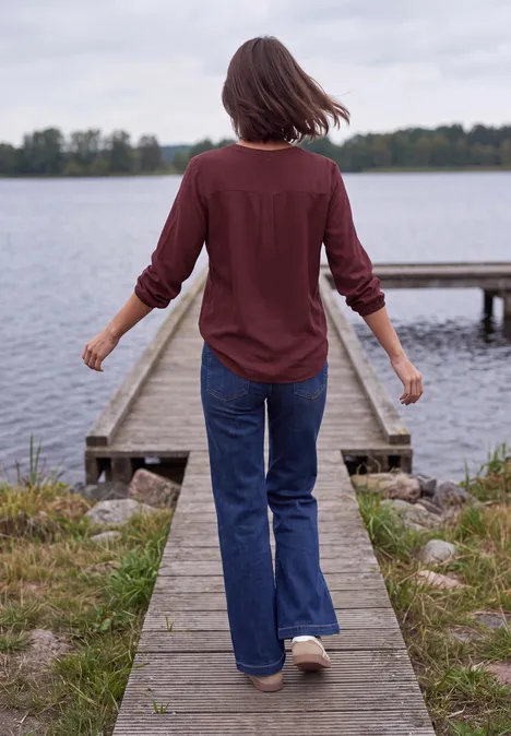 Frau in rotem Hemd und Jeans spaziert auf einem hölzernen Steg zum See, umgeben von Ufer und Bäumen.
