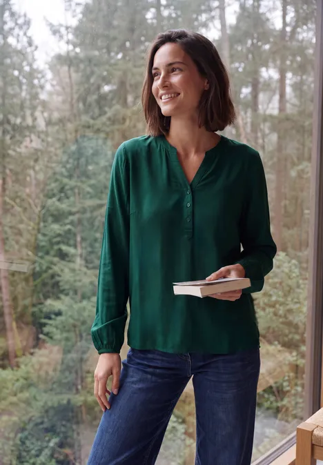 Frau in grüner Bluse steht lächelnd vor einem Waldpanorama, hält ein Buch und einen Stift in der Hand.