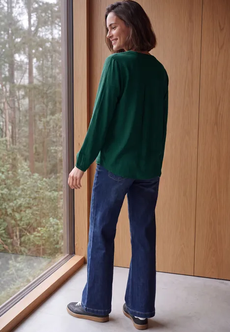 Frau steht lächelnd an großem Fenster in einem Holzraum, trägt grünen Pullover und blaue Schlagjeans.