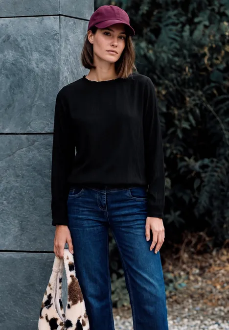 Blouse à manches longues et col rond avec détails smockés Black Femme en pull noir, jean, casquette rouge et sac, posée contre un mur à l'extérieur.