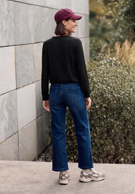 Blouse à manches longues et col rond avec détails smockés Black Personne en pull noir, jean bleu, baskets grises et casquette, debout de côté devant un mur en pierre et des buissons.