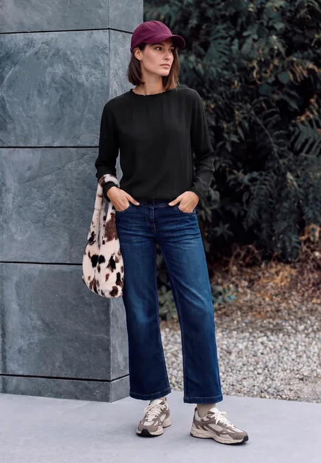 Blouse à manches longues et col rond avec détails smockés Black Femme en pull noir, jean bleu, baskets, casquette et sac, adossée contre une façade.
