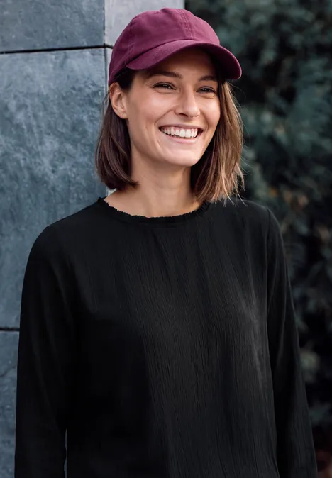 Blouse à manches longues et col rond avec détails smockés Black Une jeune femme portant une casquette bordeaux et un haut noir sourit en extérieur.