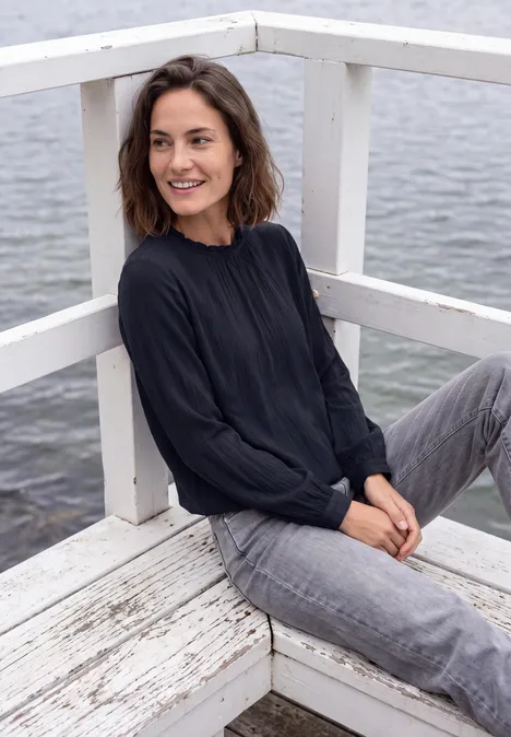 Eine Frau sitzt entspannt auf einer weißen Holzplattform am Wasser, trägt schwarzen Pullover und graue Jeans.