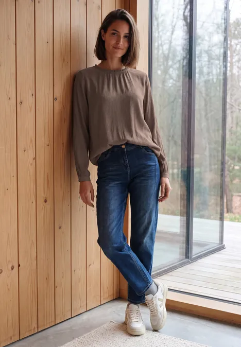 Eine junge Frau lehnt entspannt an einer Holzwand, trägt Jeans, Sneakers und ein lockeres Oberteil.