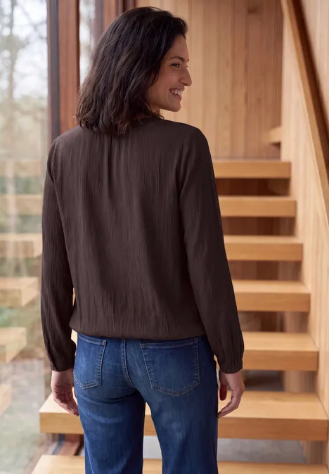 Langarm Bluse mit Rundhals und Smokdetail truffle brown Eine Frau blickt über die Schulter, trägt eine dunkle Bluse und Jeans und steht vor einer Holztreppe.