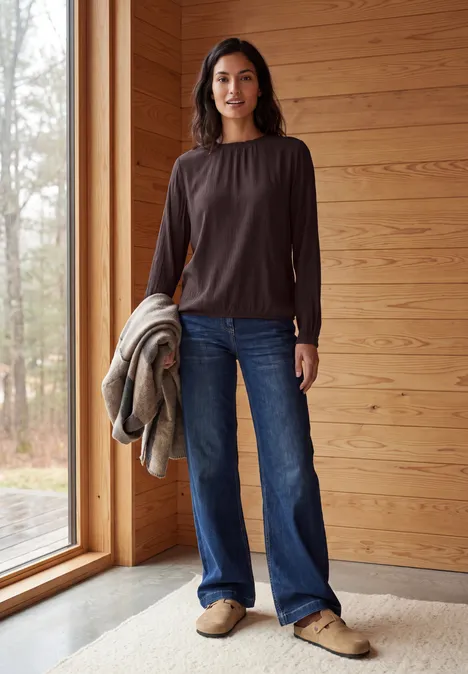 Langarm Bluse mit Rundhals und Smokdetail truffle brown Eine Frau in braunem Oberteil und Jeans steht in einem Holzhaus, hält eine Jacke, blickt in Richtung Kamera.