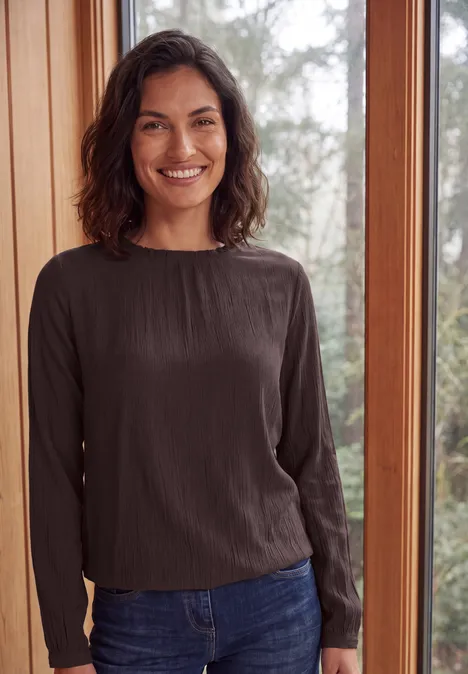 Langarm Bluse mit Rundhals und Smokdetail truffle brown Lächelnde Frau in dunkler Langarmbluse und Jeans, stehend vor großem Fenster mit Blick ins Grüne.