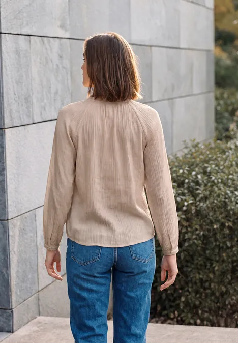 Frau mit beigem Oberteil und blauen Jeans steht mit dem Rücken zur Kamera vor Mauer und Büschen.