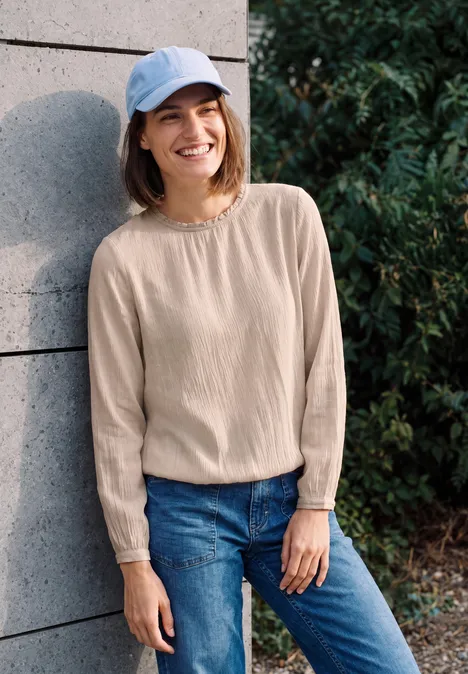 Junge Frau in blauer Kappe, beigem Pullover und Jeans lehnt draußen lächelnd an einer Wand.