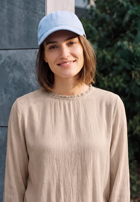 Eine Frau mit beigem Oberteil und hellblauer Baseballkappe steht lächelnd vor einer Mauer und Büschen.