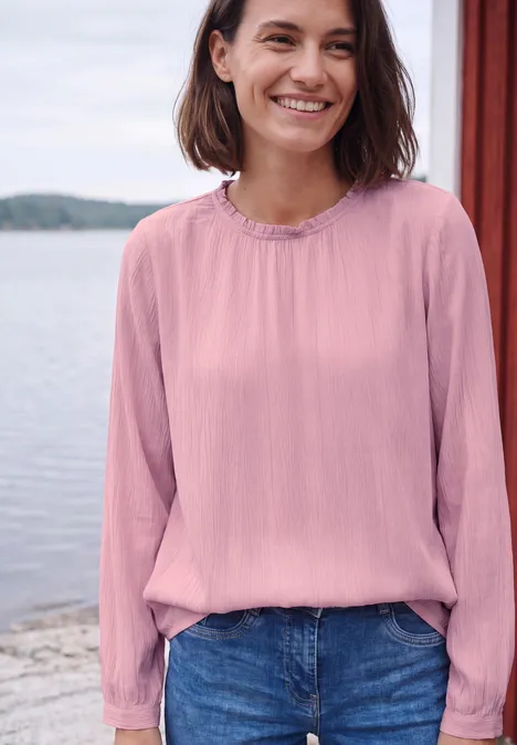 Een glimlachende vrouw in een roze blouse en spijkerbroek bij het water met houten palen op de achtergrond.