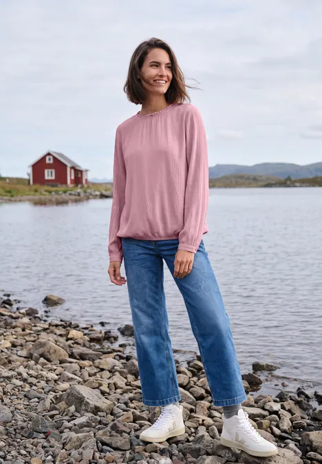 Vrouw in spijkerbroek en roze trui bij het meer, voor een rood huisje.