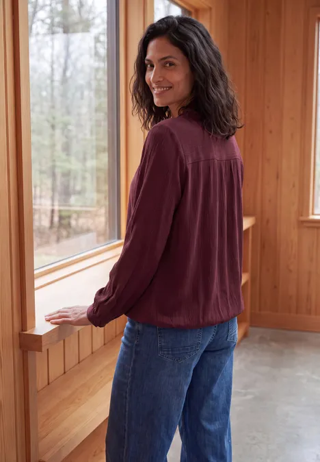 Eine lächelnde Frau mit brünetten Haaren trägt eine rote Bluse und Jeans, steht vor einem Fenster in einer Holzhütte.