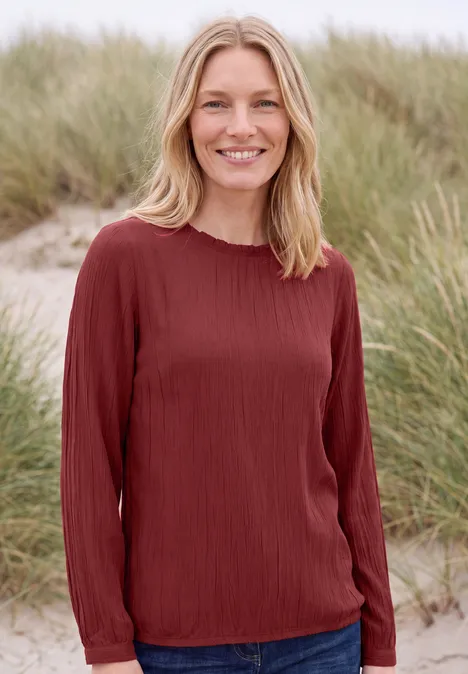 Langarm Bluse mit Rundhals und Smokdetail russet red Frau mit rotem Langarmshirt lächelt vor Dünenpflanzen