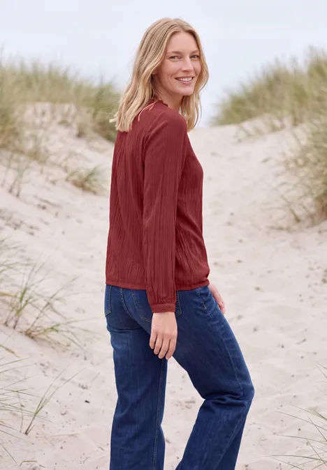 Langarm Bluse mit Rundhals und Smokdetail russet red Eine blonde Frau in rotem Pullover und Jeans steht auf Dünen am Strand und lächelt in die Kamera.