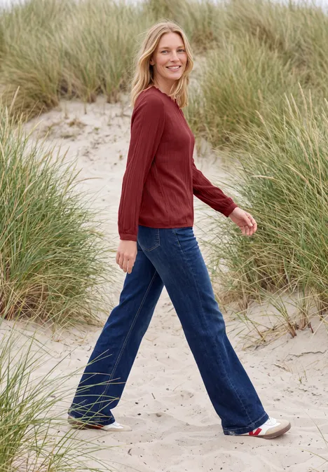 Langarm Bluse mit Rundhals und Smokdetail russet red Eine lächelnde Frau mit blonden Haaren trägt roten Pullover und blaue Jeans und geht auf sandigen Dünen mit Gras.