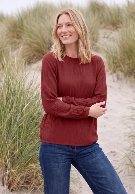 Langarm Bluse mit Rundhals und Smokdetail russet red Frau im roten Pullover und Jeans steht lächelnd am Dünenstrand.