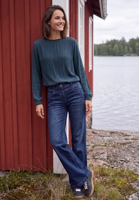 Langarm Bluse mit Rundhals und Smokdetail moonlight blue Eine lächelnde Frau in Jeans und blauem Pullover lehnt entspannt an einem roten Holzhaus am See.