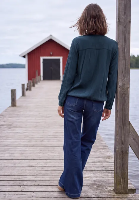 Langarm Bluse mit Rundhals und Smokdetail moonlight blue Eine Person steht auf einem Holzsteg über einem See und blickt auf ein rotes Bootshaus in der Ferne.