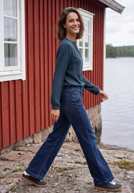 Langarm Bluse mit Rundhals und Smokdetail moonlight blue Eine Frau in Jeans und Pullover geht lächelnd an einem roten Haus am Wasser spazieren.