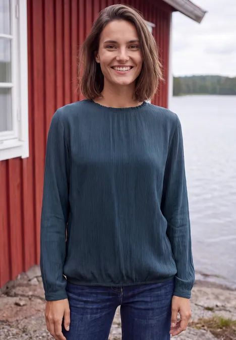 Langarm Bluse mit Rundhals und Smokdetail moonlight blue Frau mit blauem Oberteil lächelt vor rotem Haus am Wasser.