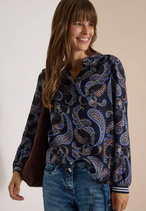 Frau mit langen braunen Haaren lächelt, trägt dunkle Paisley-Bluse und Jeans.
