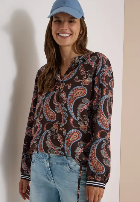 Frau mit Cap trägt Jeans und langärmlige Bluse mit buntem Paisley-Muster und lächelt.