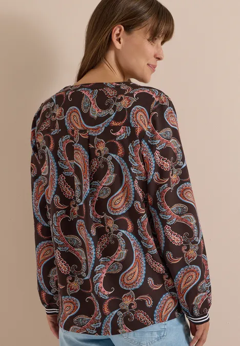 Frau von hinten in langärmliger Bluse mit buntem Paisley-Print vor hellem Hintergrund.