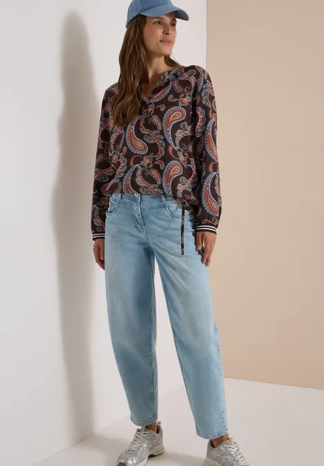 Frau in Paisley-Bluse, hellblauen Jeans, weißen Sneakern und Basecap lehnt an heller Wand.