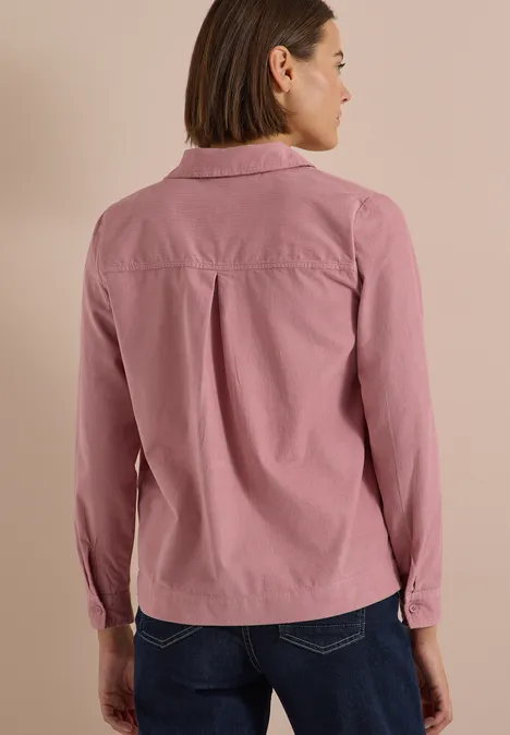 Rückansicht einer Frau mit hellrosa Hemd und Jeans vor beigem Hintergrund.