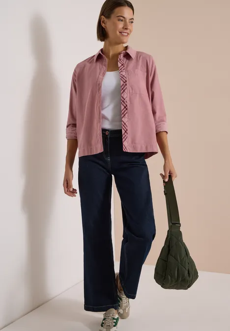 Frau trägt rosa Hemd, weißes Top, weite Jeans und Sneakers, hält grüne Handtasche vor neutralem Hintergrund.