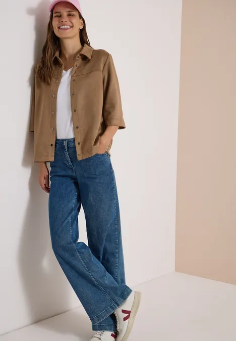 Velours Hemdbluse mit Tapedetails creamy mocha Frau in weiter Jeans, weißem Shirt, brauner Jacke und Sneakern lehnt lächelnd an der Wand.