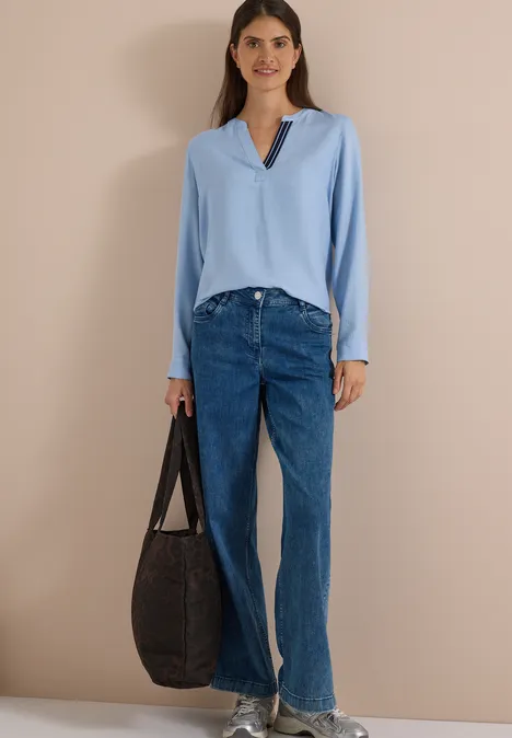 Langarm Tunika mit Split Neck und Tape blouse blue melange Frau in hellblauer Bluse, blauen Jeans, beigen Sandalen und großer dunkler Tasche vor beigem Hintergrund.