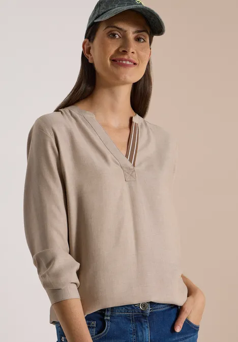 Frau in beiger Bluse, Jeans und grüner Kappe, lächelnd, mit Händen in den Hosentaschen.
