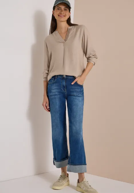 Frau vor neutralem Hintergrund in beigefarbener Bluse, umgeschlagener Jeans, Sneakern und Baseballcap.
