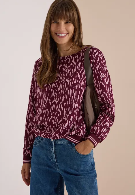 Vrouw met lang bruin haar glimlachend, gekleed in een bordeaux gemêleerde trui en jeans voor een neutrale achtergrond.