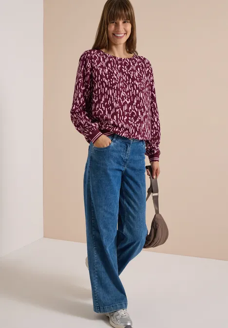 Vrouw in paarse gebloemde trui, wijde jeans en kleine schoudertas, lachend voor een neutrale achtergrond.