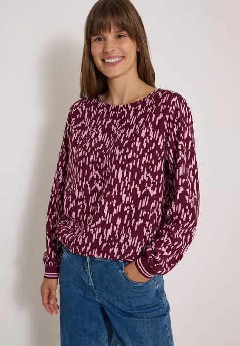 Een vrouw in een paarse blouse met dessin lacht ontspannen, met één hand in haar zak.