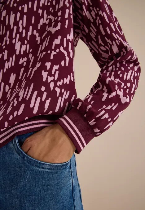 Close-up: persoon in blauwe jeans, hand in de zak, bordeauxrode trui met wit patroon.