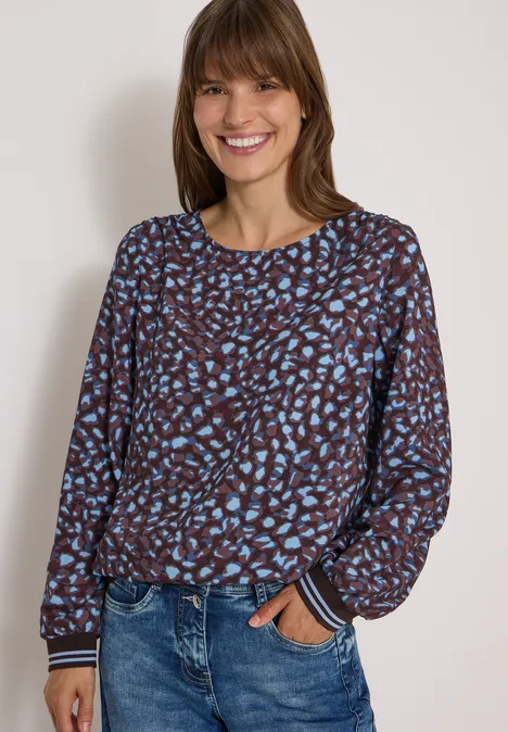 Frau lächelt, trägt braun-blau gemustertes Longsleeve und blaue Jeans vor hellem Hintergrund