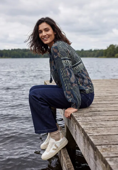 Langarm Tunika mit Bändern und Print urban dark blue Eine Frau sitzt auf einem Steg am See, trägt Jeans, gemusterte Jacke und weiße Sneaker, lächelt entspannt.