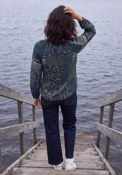 Langarm Tunika mit Bändern und Print urban dark blue Frau in gemusterter Jacke, dunklen Jeans und weißen Sneakers steht auf Holzsteg und blickt aufs Wasser.