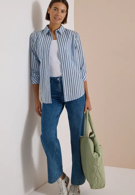 Eine Frau in gestreiftem Hemd, weißem Top, blauen Jeans und grünem Shopper lehnt an der Wand.
